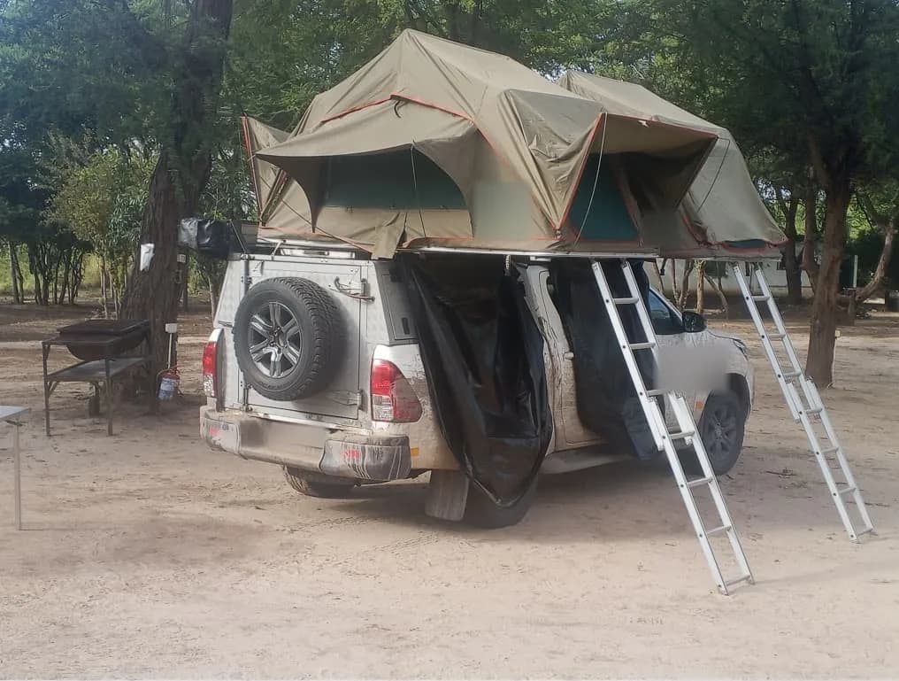 Pick-up truck with a double rooftop tent and two ladders on a shaded campsite pitch