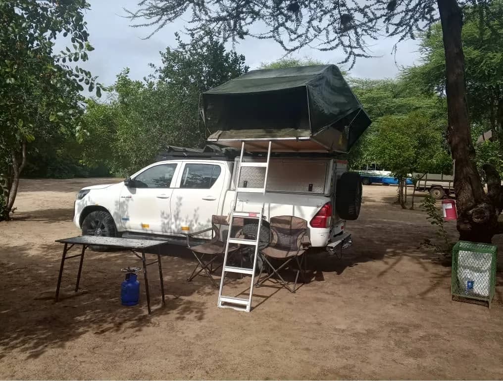 Pick-up truck with a rooftop tent and camp chairs on a shaded campsite pitch